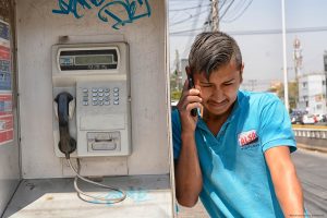 Guadalajara, Mexico - March 14 2019: Hispanic male leans on a Telmex public phone while speaking on his mobile phone in an urban setting. Modern and old obsolete communication technology