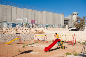 A boy plays on a bright playground in front of the security wall in the West Bank